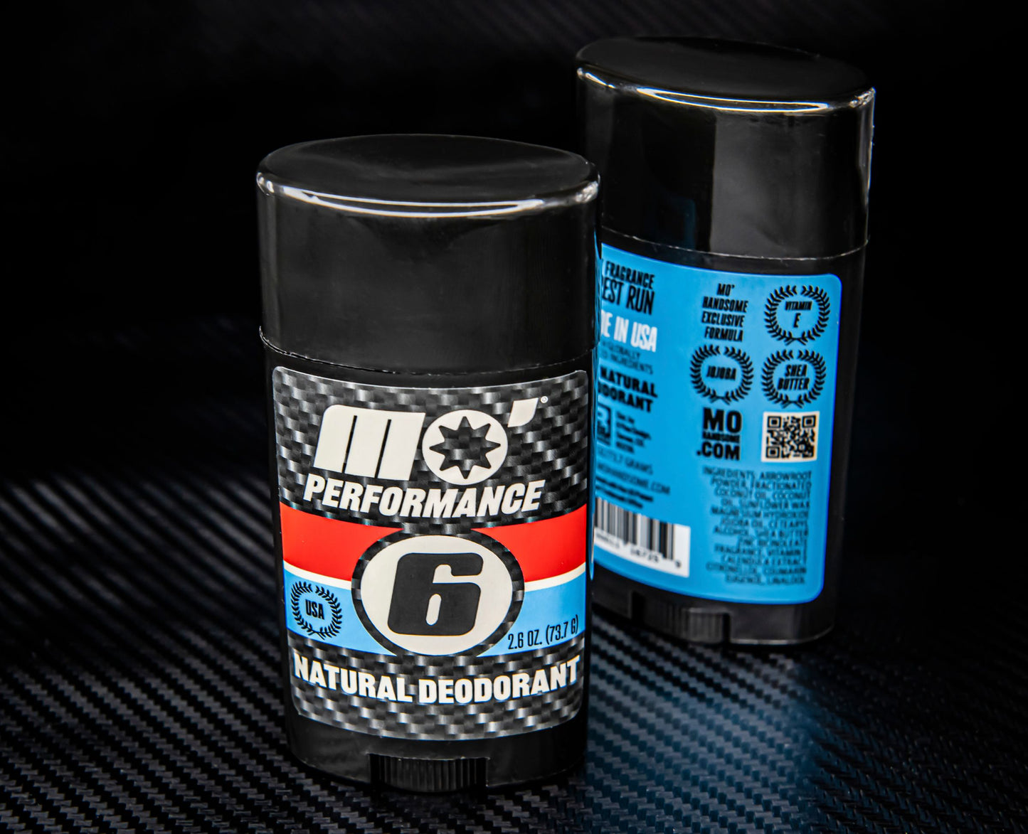 Two Mo’ Performance deodorant sticks displayed on carbon fiber surface—front branding and back label visible.
