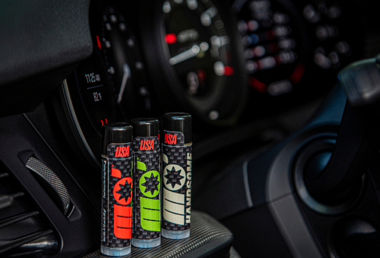 Mo’Handsome lip balm trio resting in Porsche 911 cockpit—interior carbon accents emphasize sleek, motorsport-inspired grooming.